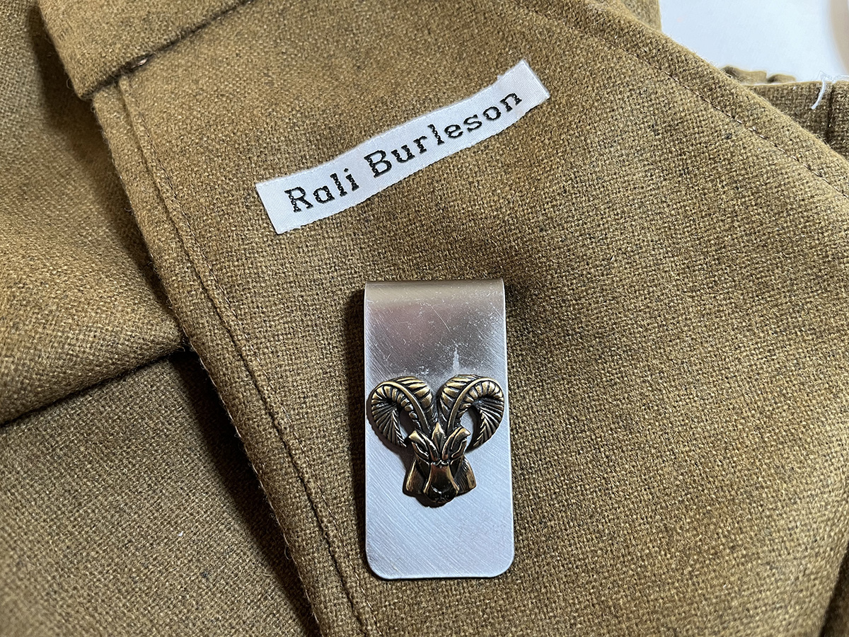 Close view of a shirt bearing a Rali Burleson name label and a ram-head metal clip. The image establishes their presence on this photographed garment.